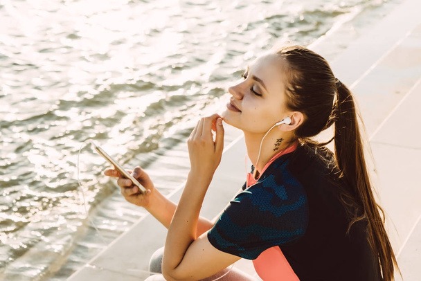 young sporty girl with long hair resting after jogging by the river, listening to music on headphones - Φωτογραφία, εικόνα