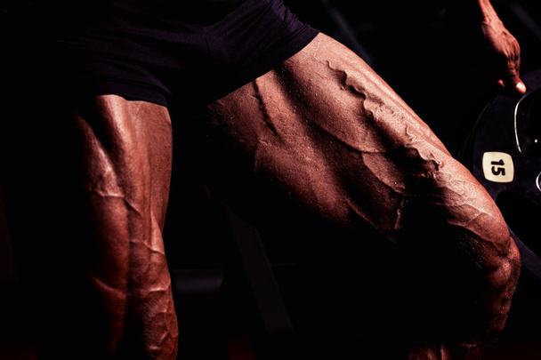Αθλητής στο γυμναστήριο. Ο τύπος κάνει ασκήσεις. Πορτραίτο closeup των ποδιών με τις φλέβες. Αρσενικό μοντέλο με μαυρισμένο δέρμα. Σέξι άντρας bodybuilder στέκεται με barbell. - Φωτογραφία, εικόνα