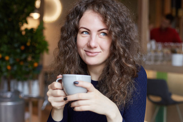 Cheerful and sleepy young attractive natural stylish female with curly hair and no make up holding big mug of coffee, enjoying fresh morning drink before lectures at university. - Photo, Image