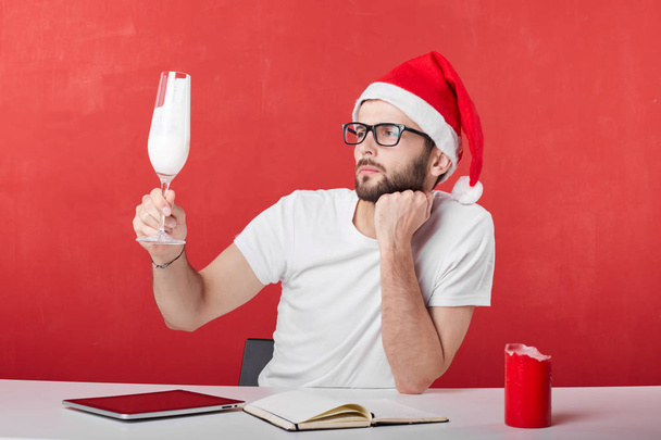 Bright and young man with interesting appearance looks with suspect at glass with white beverage, going to celebrate Christmas at work. Non alcoholic party with no people around on red wall background - Fotografie, Obrázek