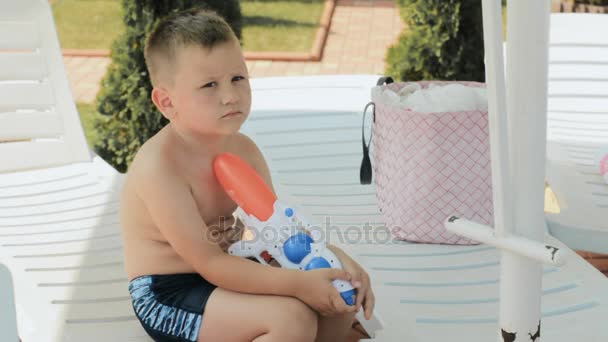 Kleiner Junge mit Wasserpistole sitzt auf einem Liegestuhl - Filmmaterial, Video