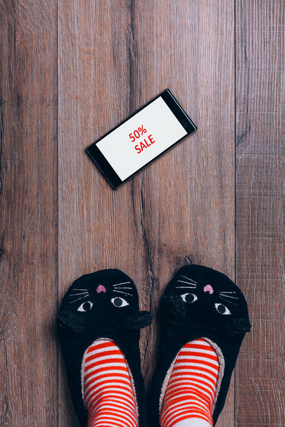 木製フローリングのスマート フォン。黒い金曜日の広告。女性足ストライプ靴下猫スリッパ。平面図です。オンライン ショッピングの概念. - 写真・画像