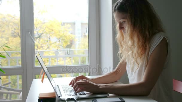 Cute young woman talking with friends via mobile phone while working at a laptop, and find helpful information to prepare for the seminar while sitting at home - Footage, Video