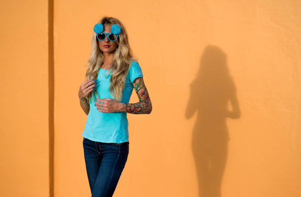 Beautiful blonde in sunglasses posing at the camera. Portrait on the background of bright orange wall. Modern hipster girl with tattoos. - Foto, Imagen