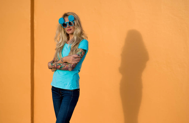Beautiful blonde in sunglasses posing at the camera. Portrait on the background of bright orange wall. Modern hipster girl with tattoos. - Fotoğraf, Görsel