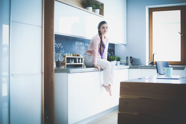 Young woman sitting on table in the kitchen. - 写真・画像