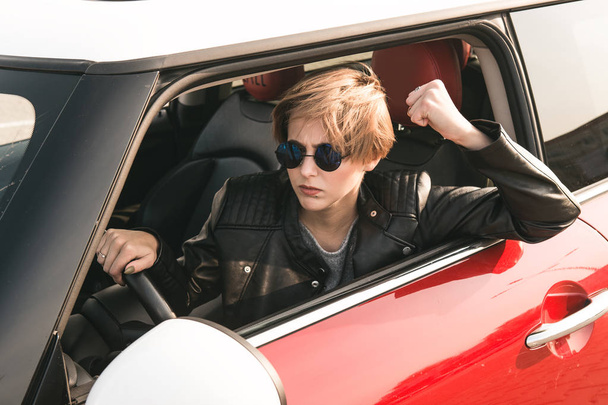 Μόδας κορίτσι οδήγηση ενός αυτοκινήτου με τα γυαλιά ηλίου. Το κορίτσι δείχνει μια γροθιά θυμωμένα και φαίνεται στην κάμερα - Φωτογραφία, εικόνα