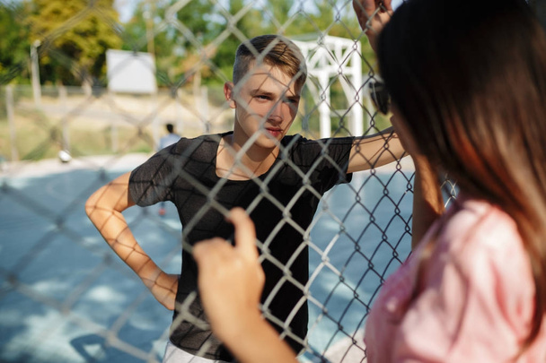 Retrato de niño bonito de pie en la cancha de baloncesto y mirando cuidadosamente a la chica con el pelo oscuro a través de valla de malla
 - Foto, Imagen
