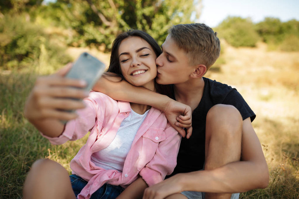 Portrait of young smiling couple sitting on lawn in park and making selfie. Beautiful couple taking photos on cellphone frontal camera - Photo, Image