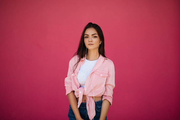 Portrait of young beautiful girl with dark hair standing and listening music in earphones while thoughtfully looking in camera on pink background isolated - Photo, Image