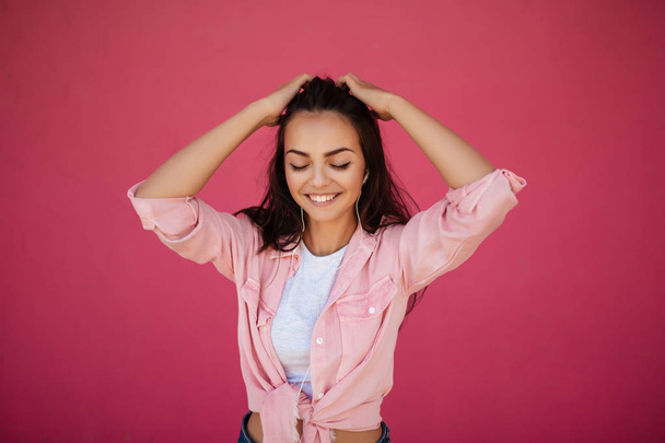 Portrait of beautiful smiling girl with dark hair standing and listening music in earphones on pink background isolated - Photo, Image