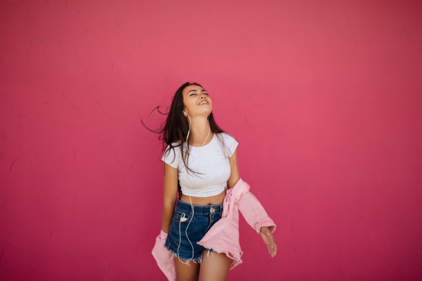 Retrato de chica alegre con el pelo largo oscuro de pie y escuchando música felizmente en auriculares sobre fondo rosa aislado
 - Foto, imagen