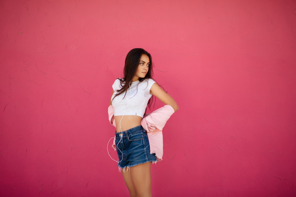 Portrait of girl with dark hair standing in denim shorts and white top while listening music in earphones and dreamily looking aside on pink background isolated - Foto, afbeelding