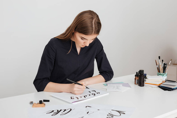 Porträt einer jungen schönen Dame, die am weißen Schreibtisch sitzt und isoliert Alphabet auf Papier schreibt - Foto, Bild