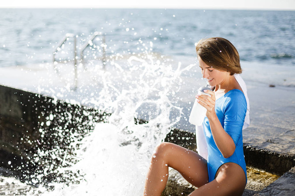 笑顔の女性が手にしうっとりと海で目を閉じて水のボトルと座っています。ブルー水着座りと背景の海の景色と水を飲んで美しい女性 - 写真・画像