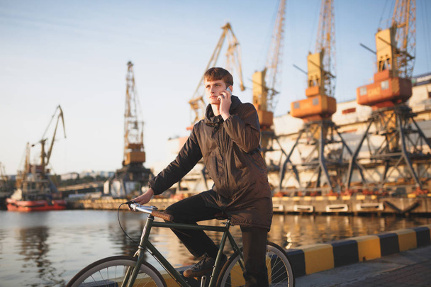 Portrait of young man with brown hair standing with bicycle and thoughtfully looking aside while talking on his mobile phone. Cool boy in down jacket on bicycle standing with sea port on background - Foto, Bild