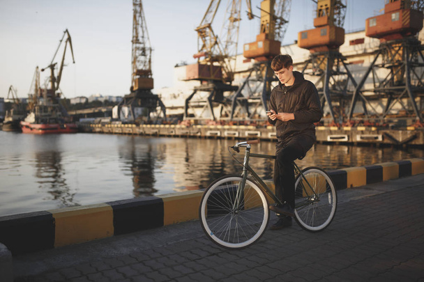 Young man with brown hair standing with bicycle and dreamily looking in his mobile phone.Thoughtful boy in down jacket on bicycle standing with sea port on background - Фото, изображение