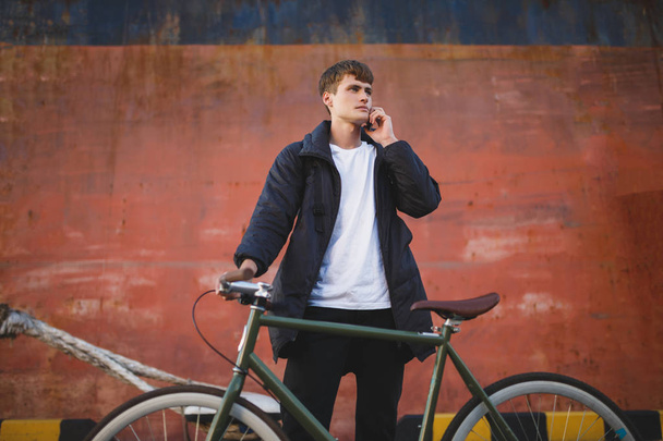 Thoughtful boy with brown hair standing with bicycle and dreamily looking aside while talking on his cellphone. Young man in down jacket standing with bicycle and mobile phone in hand - Фото, зображення
