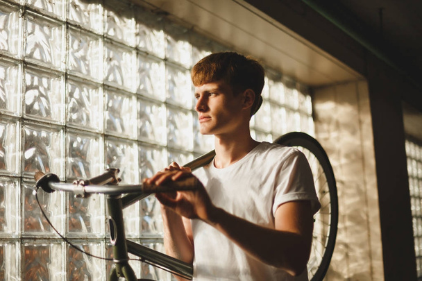 Молодой задумчивый мужчина стоит и держит велосипед, который опирается на плечо, мечтательно глядя в сторону. Портрет мальчика в белой футболке с классическим велосипедом
 - Фото, изображение