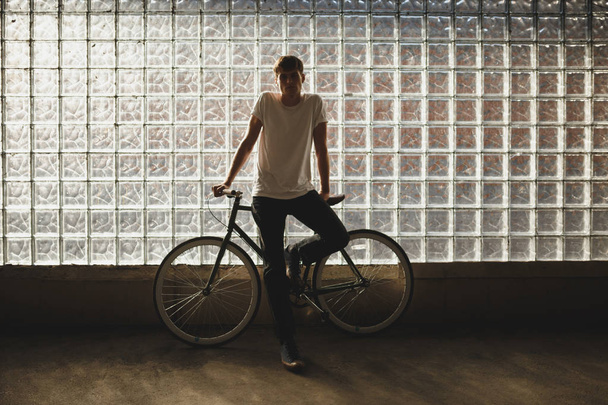Cool boy standing with classic bicycle and thoughtfully looking in camera. Photo of young man in white t-shirt leaning on bicycle with glass wall on background - Photo, Image