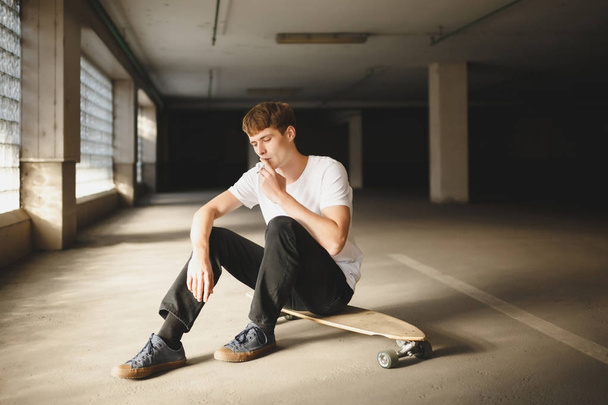 Portrait of boy with brown hair sitting on skateboard and thoughtfully looking down while smoking. Photo of young man in white t-shirt sitting and holding cigarette in hand - 写真・画像