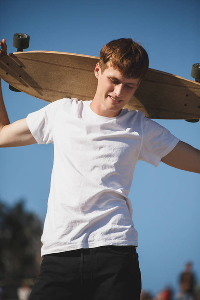 Πορτραίτο νεαρού άνδρα με καστανά μαλλιά στέκεται με skateboard στον ώμο του και απορώντας κοιτάζοντας προς τα κάτω. Νέος, χαμογελαστός άνθρωπος σε λευκό t-shirt στέκεται με skateboard στο χέρι - Φωτογραφία, εικόνα