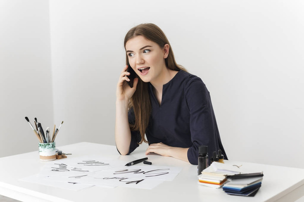 Portrait de jeune jolie dame assise au bureau blanc et parlant sur son téléphone portable tout en regardant joyeusement de côté isolé
 - Photo, image