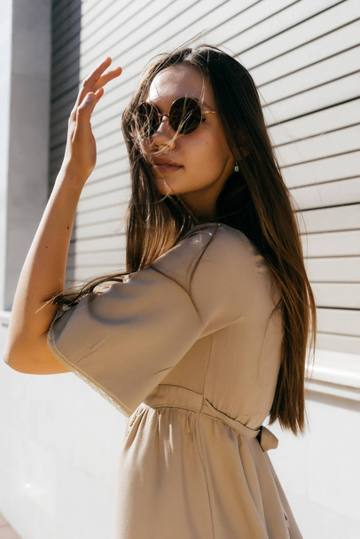 long-haired girl in a beige dress and sunglasses walking along the street in the sun - Foto, Imagen