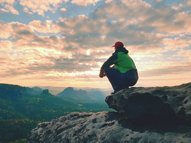 Один турист отдыхает. Турист в зеленой черной куртке, сидящий на скалистой вершине
  - Фото, изображение