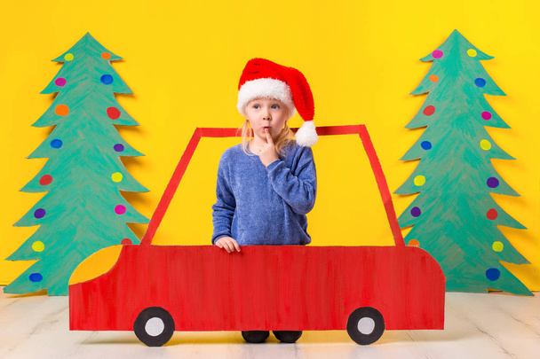 Kind mit Weihnachtsmütze am Steuer eines Autos aus Pappe. Weihnachtskonzept. Neujahrsferien. - Foto, Bild