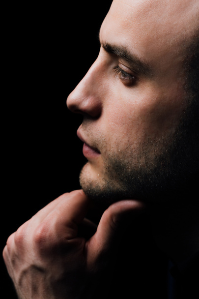 Studio portrait of young sexy man in black shirt, holding hands in chin, looking down with humility. - Photo, Image