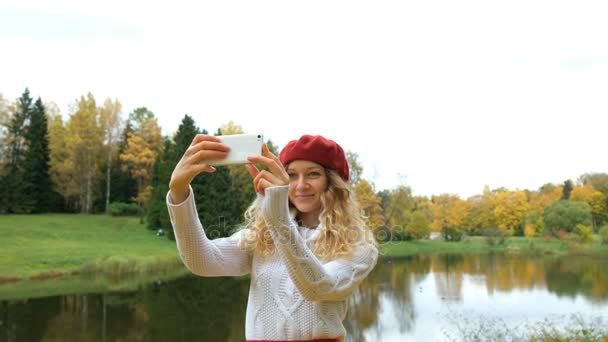 Charmante jonge Kaukasische vrouw in een little red riding hood poseren en het nemen van een selfie op uw moderne smartphone en glimlachend in het najaar van Park. - Video