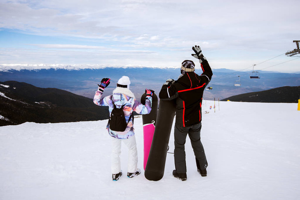 Achteraanzicht van gelukkige paar snowboards te houden en met de handen omhoog genieten in de winter van de besneeuwde bovenop berg - Foto, afbeelding