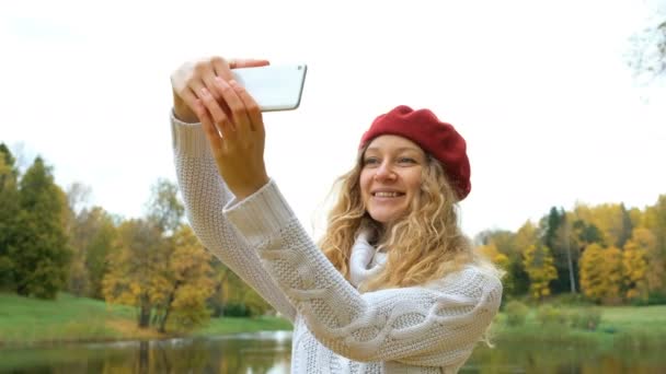 Encantadora joven mujer caucásica en una pequeña capucha roja posando y tomando una selfie en su teléfono inteligente moderno y sonriendo en otoño Park
. - Imágenes, Vídeo