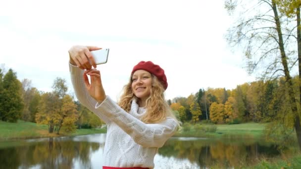 Encantadora joven mujer caucásica en una pequeña capucha roja posando y tomando una selfie en su teléfono inteligente moderno y sonriendo en otoño Park
. - Imágenes, Vídeo