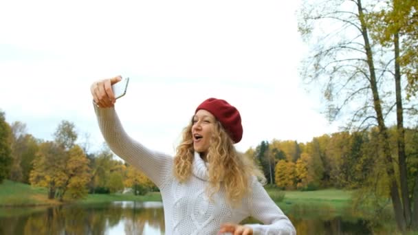 Charmante junge kaukasische Frau mit Rotkäppchen posiert und macht ein Selfie mit Ihrem modernen Smartphone und lächelt im Herbstpark. - Filmmaterial, Video