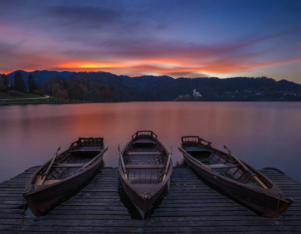 Bled, Słowenia - tradycyjne łodzie w jezioro Bled z zachodzące dramatyczna i Wniebowzięcie Maryi kościół pielgrzymkowy i górami w tle o zachodzie słońca  - Zdjęcie, obraz