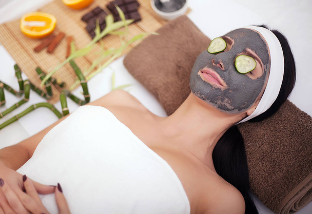 Beautiful young woman is getting facial clay mask at spa, lying with cucumbers on eyes - Фото, зображення