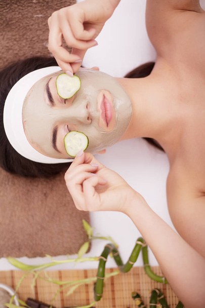 Beautiful young Woman in clay mud mask on face covering eyes with slices cucumber. - Photo, Image