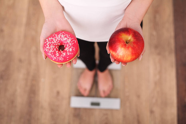 Dieta. Donna che misura il peso corporeo sulla bilancia tenendo ciambella e mela. I dolci sono cibo spazzatura malsano. Dieta, Mangiare sano, Stile di vita. Perdita di peso. Obesità. Vista dall'alto
 - Foto, immagini