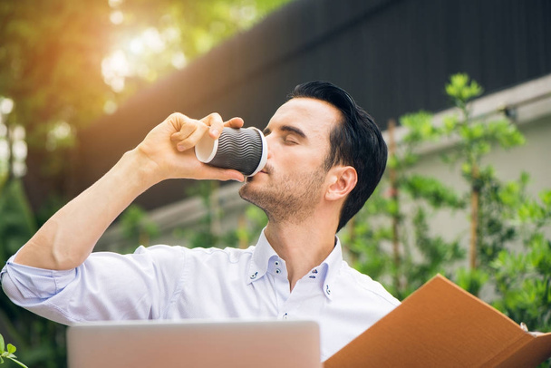 Portret przystojny człowieka sukcesu picia kawy. Szczęśliwy człowiek wziąć przerwę na kawę jak on siedzi na swoim laptopie działa z domowym biurze z regału. działalności człowieka o śniadanie siedzący odkryty - Zdjęcie, obraz