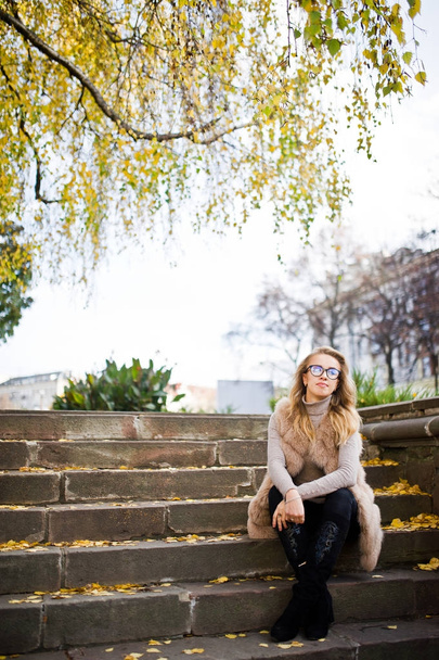 Blonde girl at fur coat and glasses sitting on stairs with yello - Φωτογραφία, εικόνα