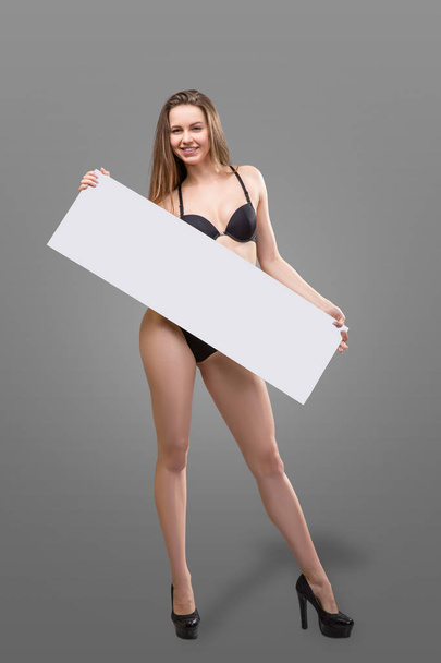 Jovem mulher em lingerie preta com sinal em branco na frente de fundo cinza
 - Foto, Imagem