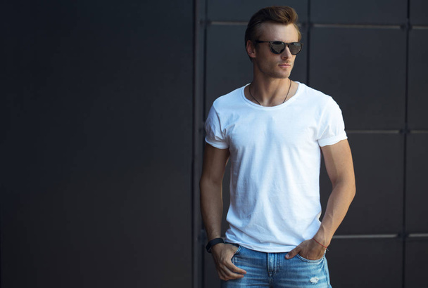 Street fashion concept. Portrait of a young and handsome man in a white t shirt and trendy glasses posing over black modern background. Close up. Street shot. copy space - Фото, зображення