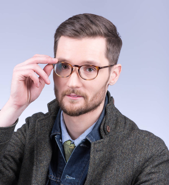 A young, stylish man with a beard adjusts his glasses. Modern dress, studio portrait - Фото, изображение
