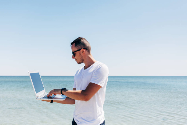 Молодой человек держит белый ноутбук на фоне моря
 - Фото, изображение