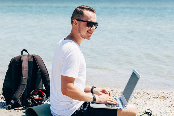 Mladý muž v bílé tričko sedí na pláži s notebookem na pozadí moře - Fotografie, Obrázek