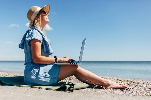Młoda kobieta na sobie niebieską sukienkę, siedząc na plaży z laptopa w jej ramiona, na tle morza - Zdjęcie, obraz