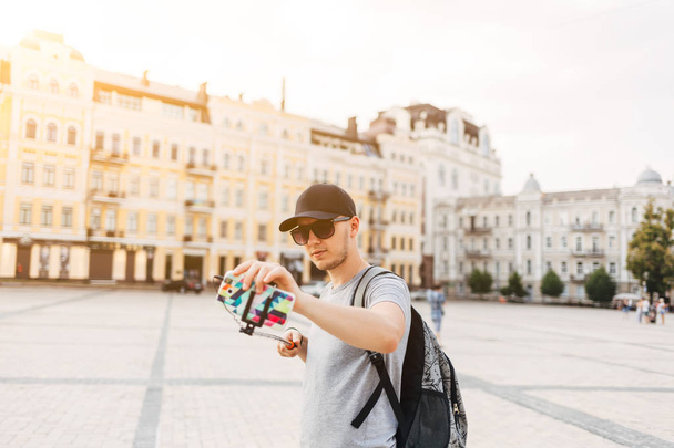 Joven alegre en camisa gris y gorra negra con mochila hacer selfie en el teléfono en Kiev Ucrania. Hombre fotografiándose con monópodo
 - Foto, Imagen