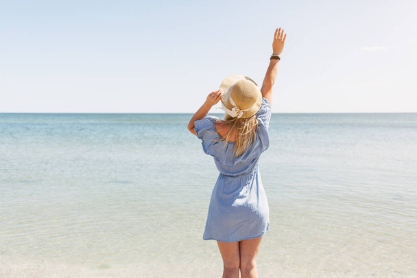 Mooie jonge gelukkige vrouw in een strooien hoed en een blauwe jurk in de achtergrond van de zee. Achteraanzicht - Foto, afbeelding